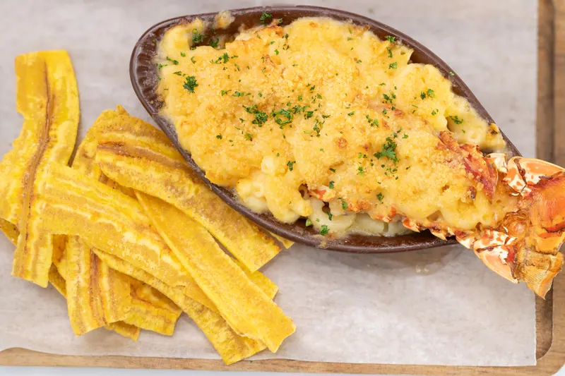 Lobster macaroni and cheese served on a wooden board with a stack of golden fried plantain strips